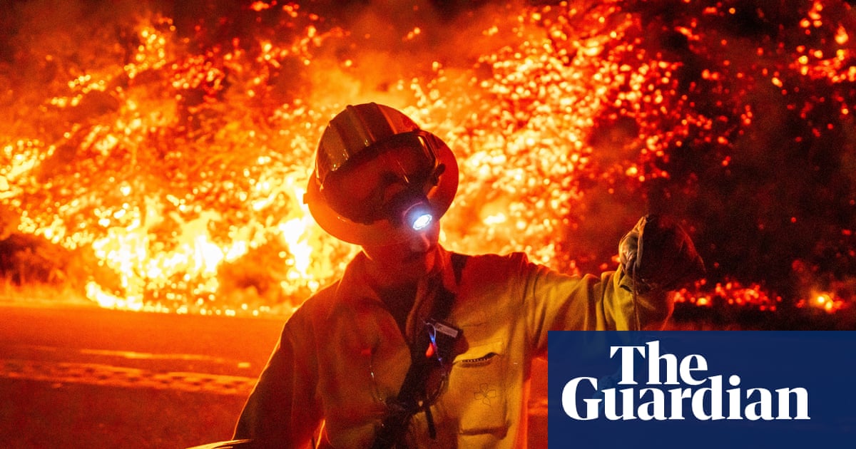 A person in a yellow jacket, helmet with goggles, holds a rake in one hand and the tail of a small rodent with the other as a verge behind him, across the street, burns with orange flames.