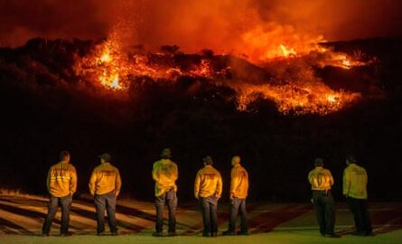 A line of seven people in yellow reflective jackets, lit from behind, watch orange flames on a black hillside.