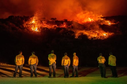 A line of seven people in yellow reflective jackets, lit from behind, watch orange flames on a black hillside.