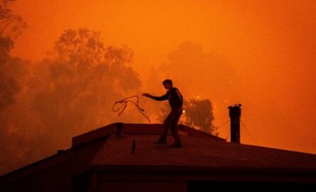 Silhouette of man on roof of house, with sky completely orange and smoky.