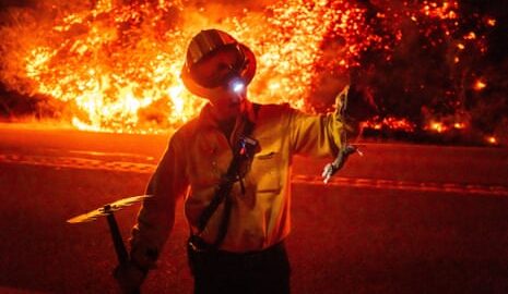A person in a yellow jacket, helmet with goggles, holds a rake in one hand and the tail of a small rodent with the other as a verge behind him, across the street, burns with orange flames.