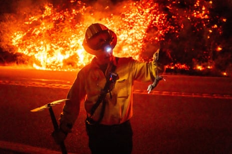 A person in a yellow jacket, helmet with goggles, holds a rake in one hand and the tail of a small rodent with the other as a verge behind him, across the street, burns with orange flames.