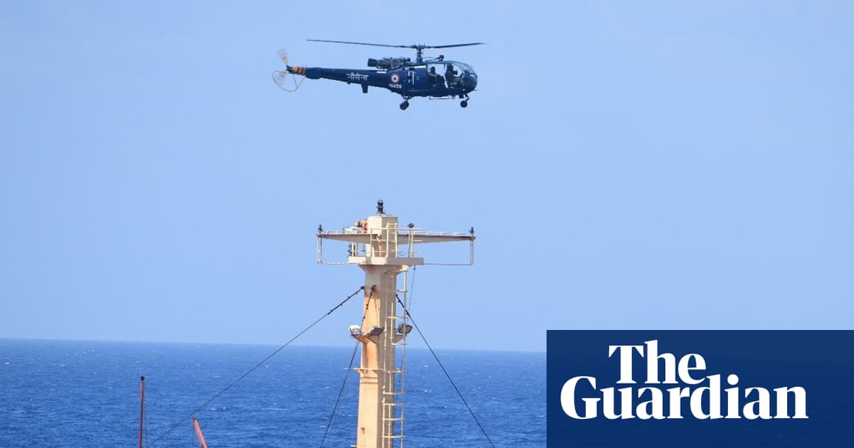 A helicopter hovers above a large bulk carrier vessel with a black-painted hull as people huddle on the deck.