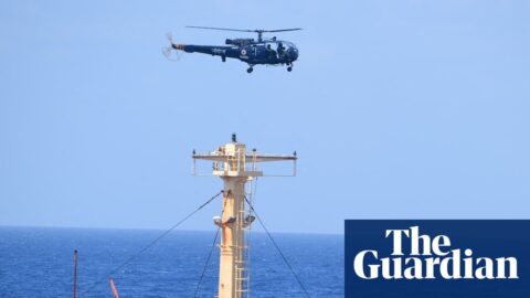 A helicopter hovers above a large bulk carrier vessel with a black-painted hull as people huddle on the deck.