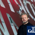 Michael Sheen stands before a large red and white lettered backdrop at the Wales Millennium Centre