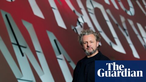 Michael Sheen stands before a large red and white lettered backdrop at the Wales Millennium Centre