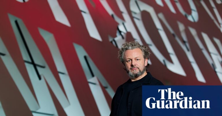 Michael Sheen stands before a large red and white lettered backdrop at the Wales Millennium Centre