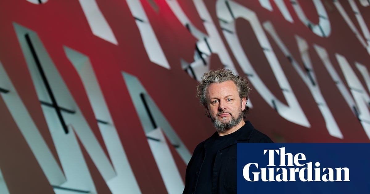 Michael Sheen stands before a large red and white lettered backdrop at the Wales Millennium Centre