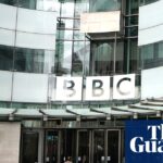 The curved frontage of BBC Broadcasting House in Portland Place, London, with the BBC logo on large glass windows above the doors. People with cameras stand in the courtyard in the foreground with others talking and going towards the doors.