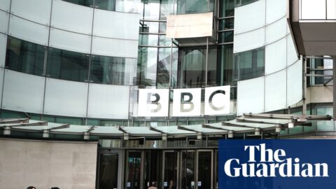 The curved frontage of BBC Broadcasting House in Portland Place, London, with the BBC logo on large glass windows above the doors. People with cameras stand in the courtyard in the foreground with others talking and going towards the doors.