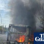 Wreckage of burning bus with city street and buildings beyond