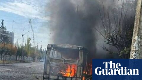 Wreckage of burning bus with city street and buildings beyond