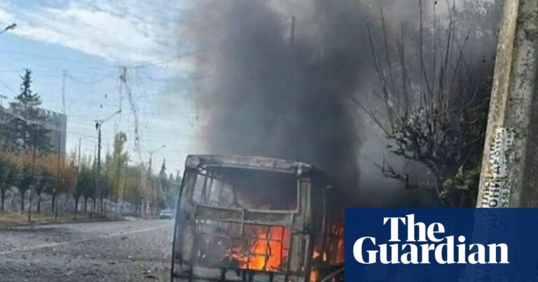 Wreckage of burning bus with city street and buildings beyond