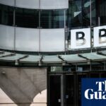 A man walks across a sunlit plaza beneath a BBC sign at Broadcasting House