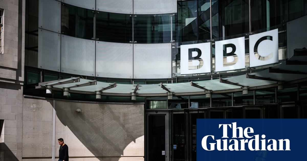 A man walks across a sunlit plaza beneath a BBC sign at Broadcasting House